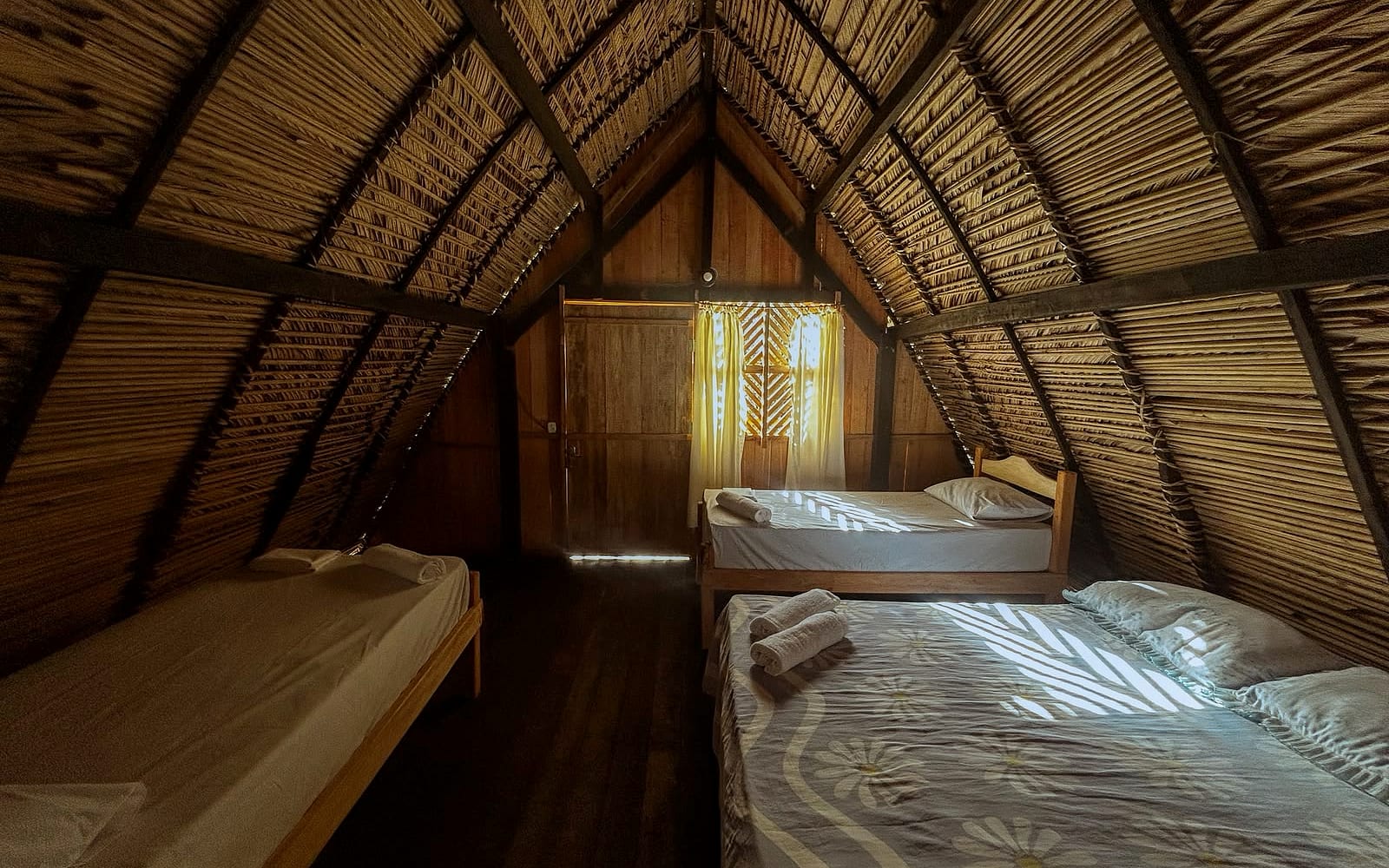 Vista ampla do quarto com cama de casal e cama de solteiro sob o telhado de palha